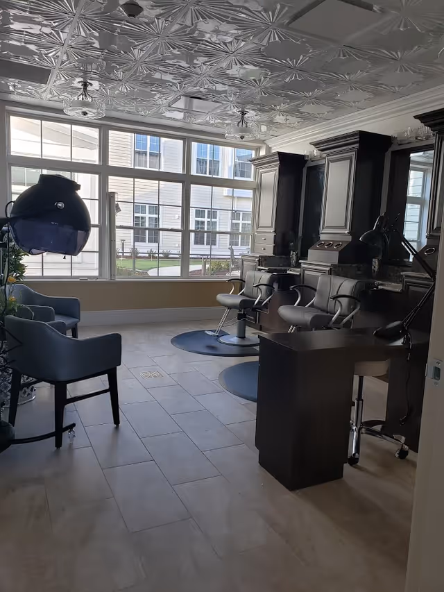 Interior of a salon area with large windows letting in natural light. The room features salon chairs, hair dryers, and dark wood cabinetry with mirrors. The ceiling has a decorative pattern and the floor is tiled.