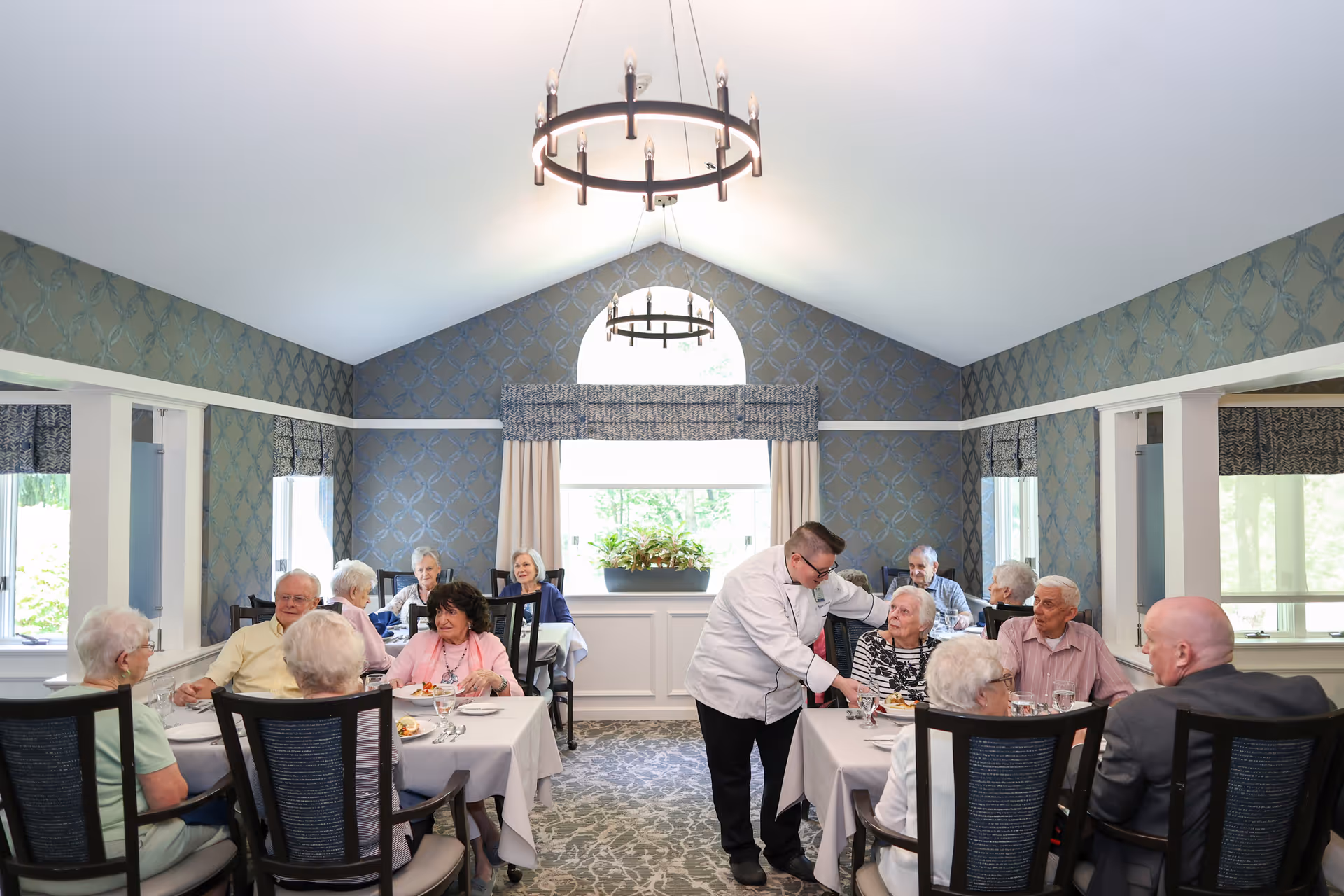 A dining room in a senior living facility with elderly residents seated at tables covered with white tablecloths. A staff member in a white uniform is serving food to one of the residents. The room has patterned wallpaper, large windows with curtains, and two circular chandeliers hanging from a vaulted ceiling.