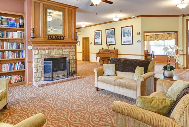 A cozy living room area in a senior living facility featuring a stone fireplace with a wooden mantle and a mirror above it. There are bookshelves filled with books on the left side, a beige sofa and armchairs with cushions, a wooden piano against the far wall, and framed pictures hanging above it. The room has patterned carpet flooring and warm lighting with ceiling fans and light fixtures.