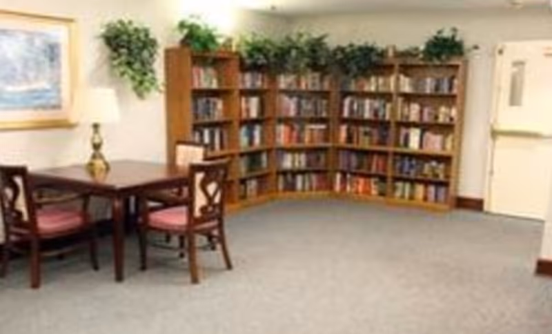 A cozy common room with a wooden table and chairs, a corner bookshelf filled with books, and greenery on top.