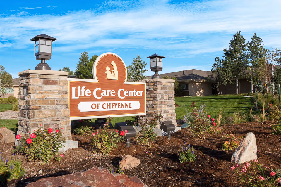Outdoor view of the Life Care Center of Cheyenne sign surrounded by landscaped garden with flowers, rocks, and two stone pillars with lanterns. A building and trees are visible in the background under a blue sky with some clouds.