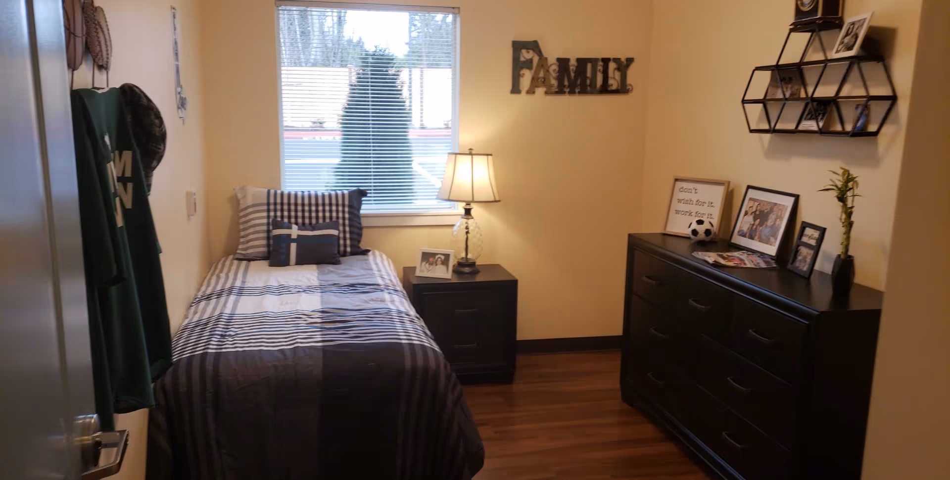 Small single bedroom with a twin bed, nightstand and lamp beneath a window, and a dresser with photos and wall decor reading 'Family'.