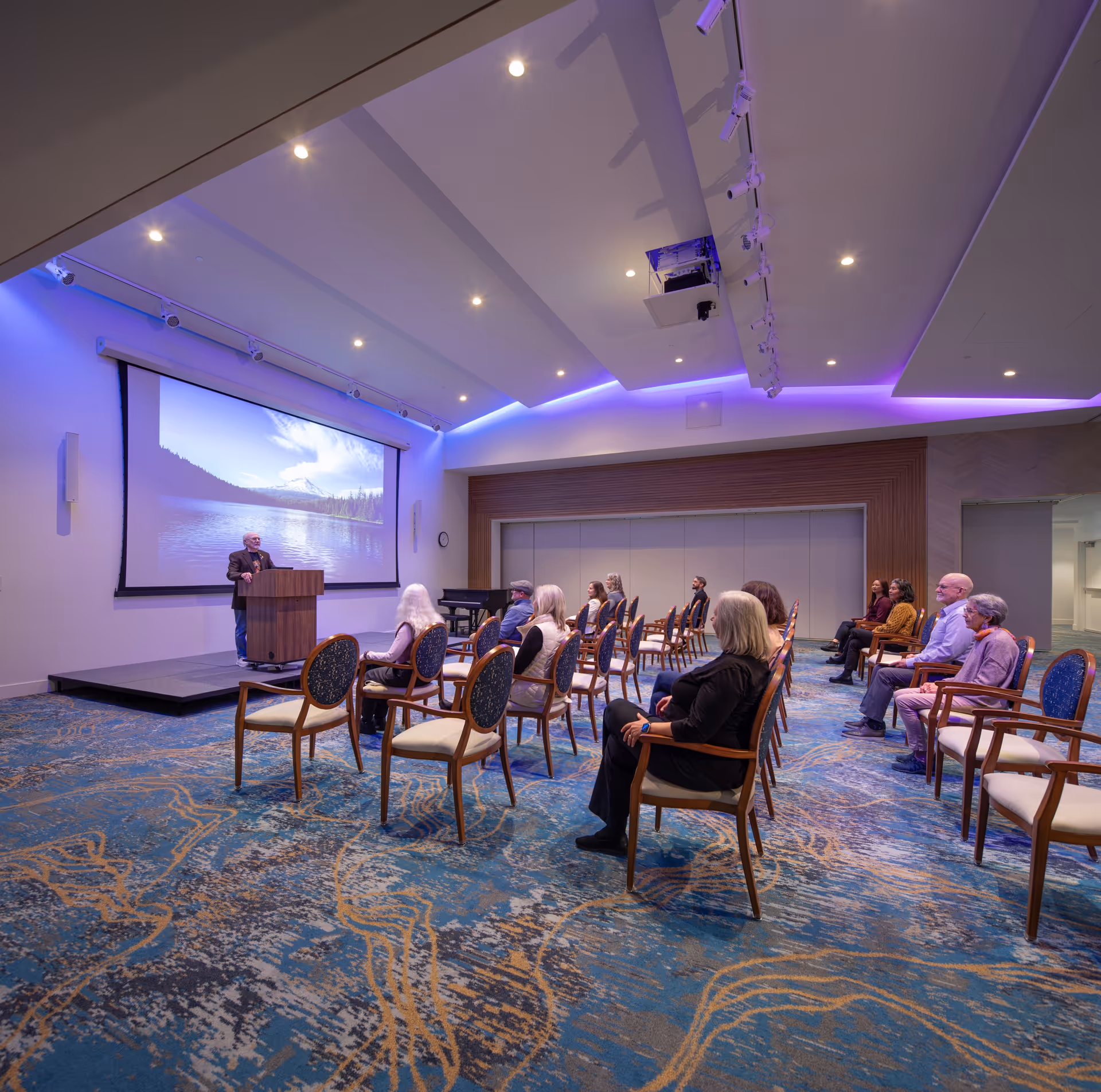 A group of elderly people seated in a spacious room with blue and gold patterned carpet, facing a man speaking at a podium in front of a large projection screen displaying a scenic lake and mountain image. The room has modern lighting with purple accent lights along the ceiling.