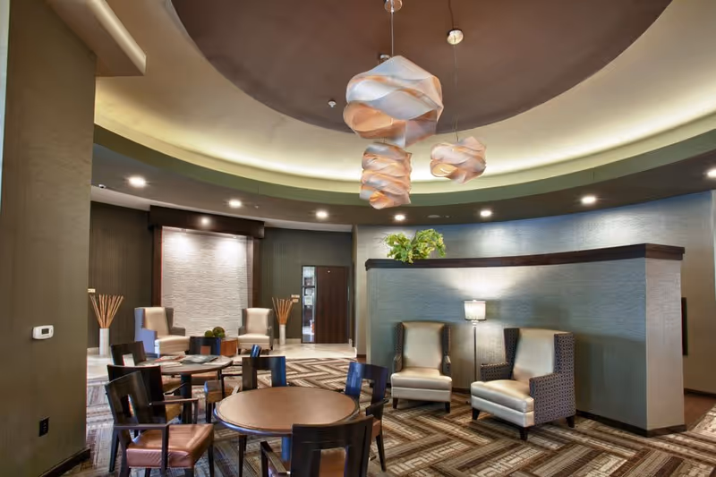 A modern senior living facility common area with a circular ceiling design featuring three artistic pendant lights. The room has a mix of seating including round tables with chairs and two armchairs near a half wall with a lamp and a plant on top. The floor is carpeted with a geometric pattern, and the walls are decorated with textured panels and neutral tones.