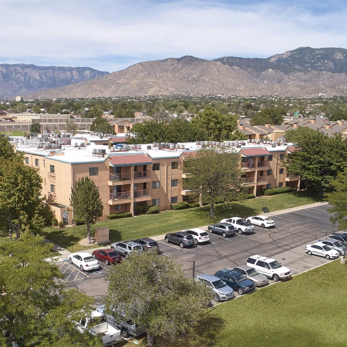 Aerial view of Sandia Vista Senior Living facility showing multiple beige apartment buildings with red-tiled awnings over balconies, surrounded by green trees and a parking lot filled with cars. In the background, there are mountains under a partly cloudy sky.