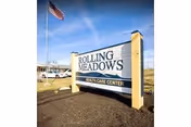 Outdoor view of a large sign for Rolling Meadows Health Care Center with an American flag flying on a flagpole nearby under a clear blue sky.