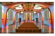 Interior view of a chapel with wooden pews arranged in rows facing an altar at the front. The ceiling features wooden beams and modern light fixtures. Stained glass windows line the sides, casting colorful light into the space. A large cross is mounted on the wall behind the altar.