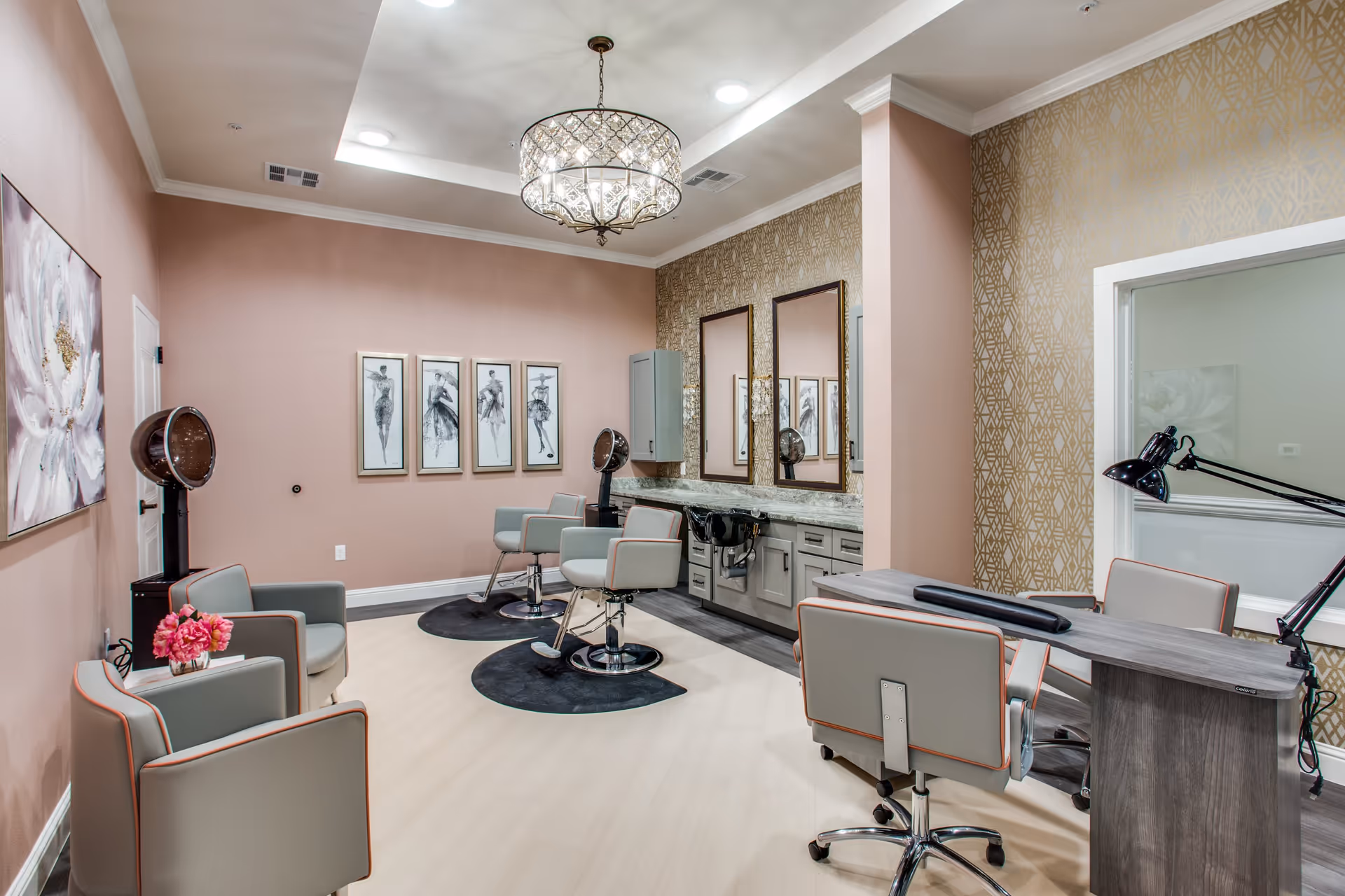 Interior view of a senior living facility's salon area with two hair styling chairs in front of mirrors and sinks, two hair dryers, a manicure table with two chairs and lamps, and decorative artwork on the walls. The room features soft pink walls, patterned wallpaper, and a chandelier ceiling light.
