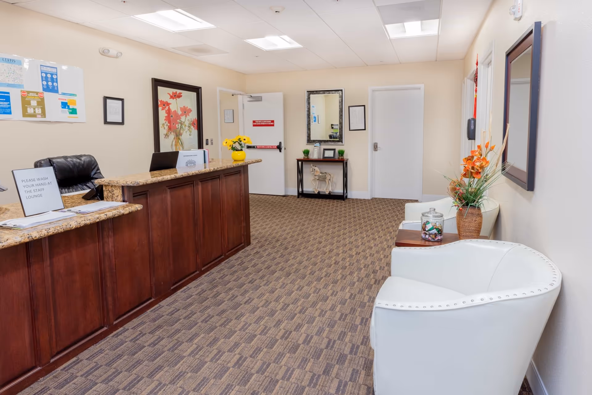 Reception area of Milan Villa Senior Living with a wooden front desk, a black office chair, a sign asking to wash hands at the staff lounge, a vase with yellow flowers on the counter, a framed floral painting on the wall, two white chairs with a small table holding a jar of candy and a vase with orange flowers, a mirror above a small table with decorative items, and beige walls and carpeted floor.