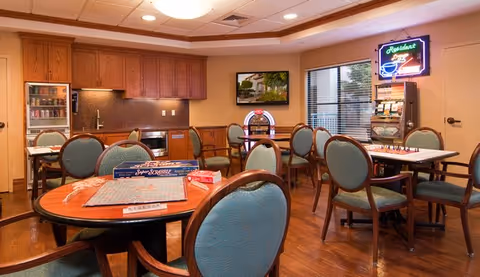 A cozy common area with several round tables and green cushioned chairs. One table has a Scrabble game set up. The room features wooden cabinetry with a small refrigerator, microwave, and sink. A neon sign and a slot machine are visible near the window, and a flat-screen TV is mounted on the wall.