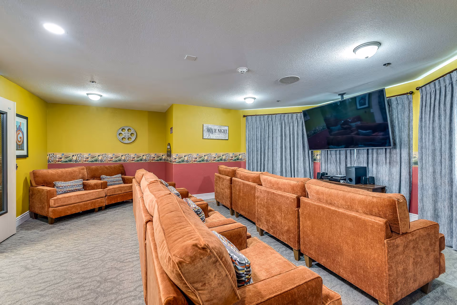 A cozy senior living facility lounge with multiple orange upholstered sofas arranged in rows facing a large wall-mounted flat screen TV. The walls are painted yellow with a decorative border featuring movie reels and film motifs. There are framed pictures and a sign that reads 'Movie Night' on the walls. The room is carpeted and has ceiling lights providing illumination.