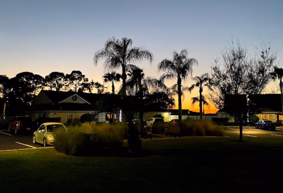 Sunset view of Palm Cottages of Rockledge with palm trees, parked cars, benches, and a building in the background under a clear sky.