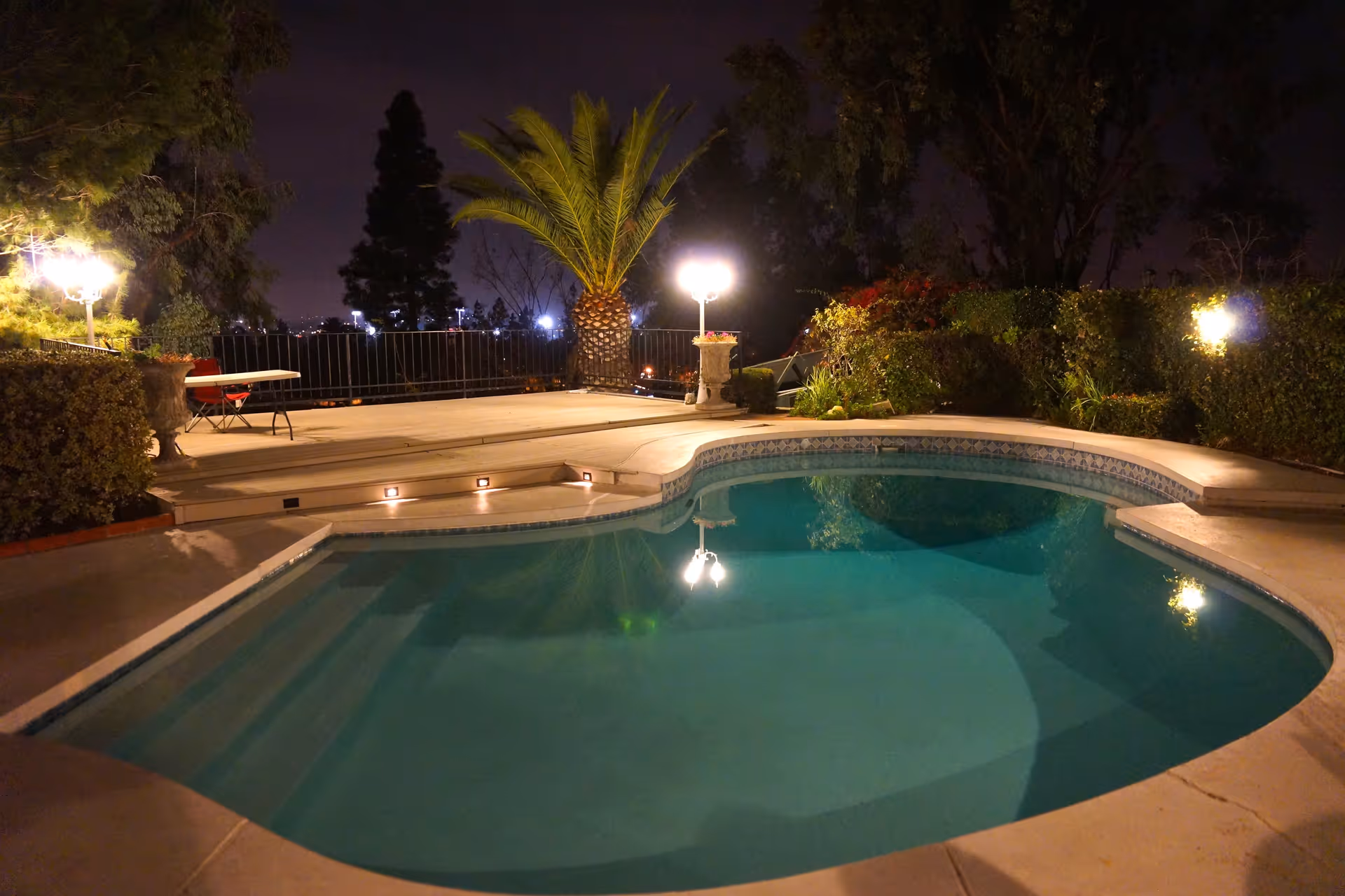 Nighttime view of a swimming pool surrounded by a concrete deck with steps leading up to a raised patio area. The patio has a table and chairs, and there are palm trees, bushes, and other greenery around the pool area. Several outdoor lights illuminate the scene.