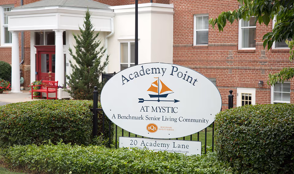 Outdoor view of the entrance to Academy Point at Mystic, a senior living community, showing a white oval sign with the community name and logo in front of a brick building with white columns and red doors, surrounded by green bushes and trees.