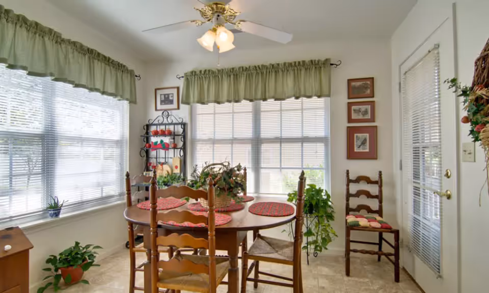 Sunlit dining nook with a round wooden table and chairs, placemats, plants and large windows with valances.