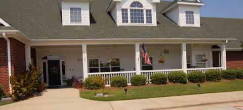Front exterior of Sumter Terrace Assisted Living with a porch, flag, and green shrubs.