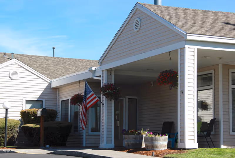 Front entrance of a beige single-story assisted living building with an American flag, hanging flower baskets, and barrel planters.