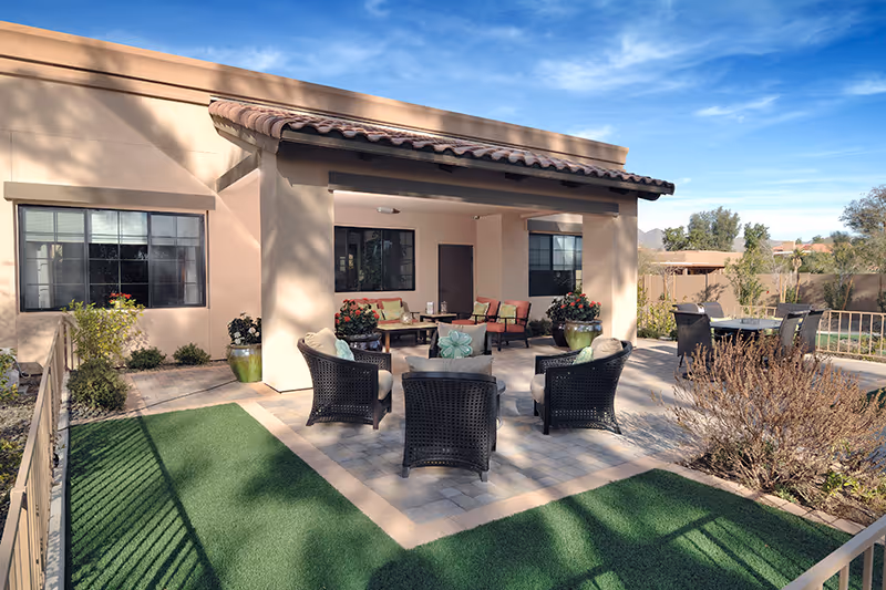 Sunlit outdoor patio with wicker seating, dining table, potted plants, and artificial turf in front of a stucco building.