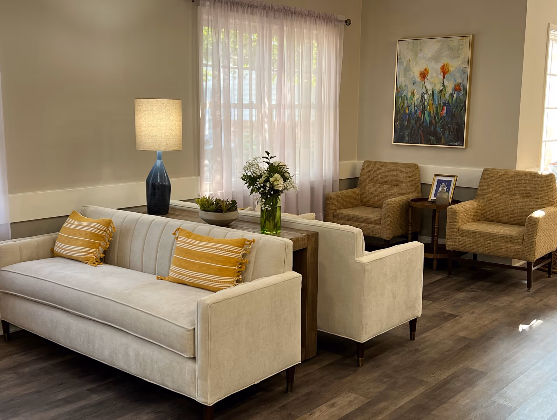 A cozy living room area with a beige sofa adorned with two yellow striped pillows, a wooden side table with a blue lamp, a green vase with white flowers, two beige armchairs, a small round wooden table with a framed photo and a decorative item, a large window with sheer white curtains, and a colorful floral painting on the wall.