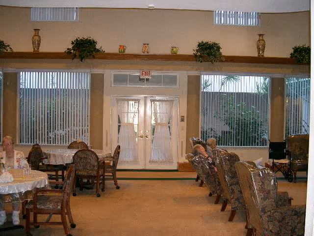 Interior view of a senior living facility common area with multiple tables and chairs. Two elderly women are seated, one at a round table on the left and another in a row of armchairs on the right. The room has large windows with vertical blinds, a set of double doors with sheer curtains, and decorative plants and vases on a high shelf above the doors and windows.
