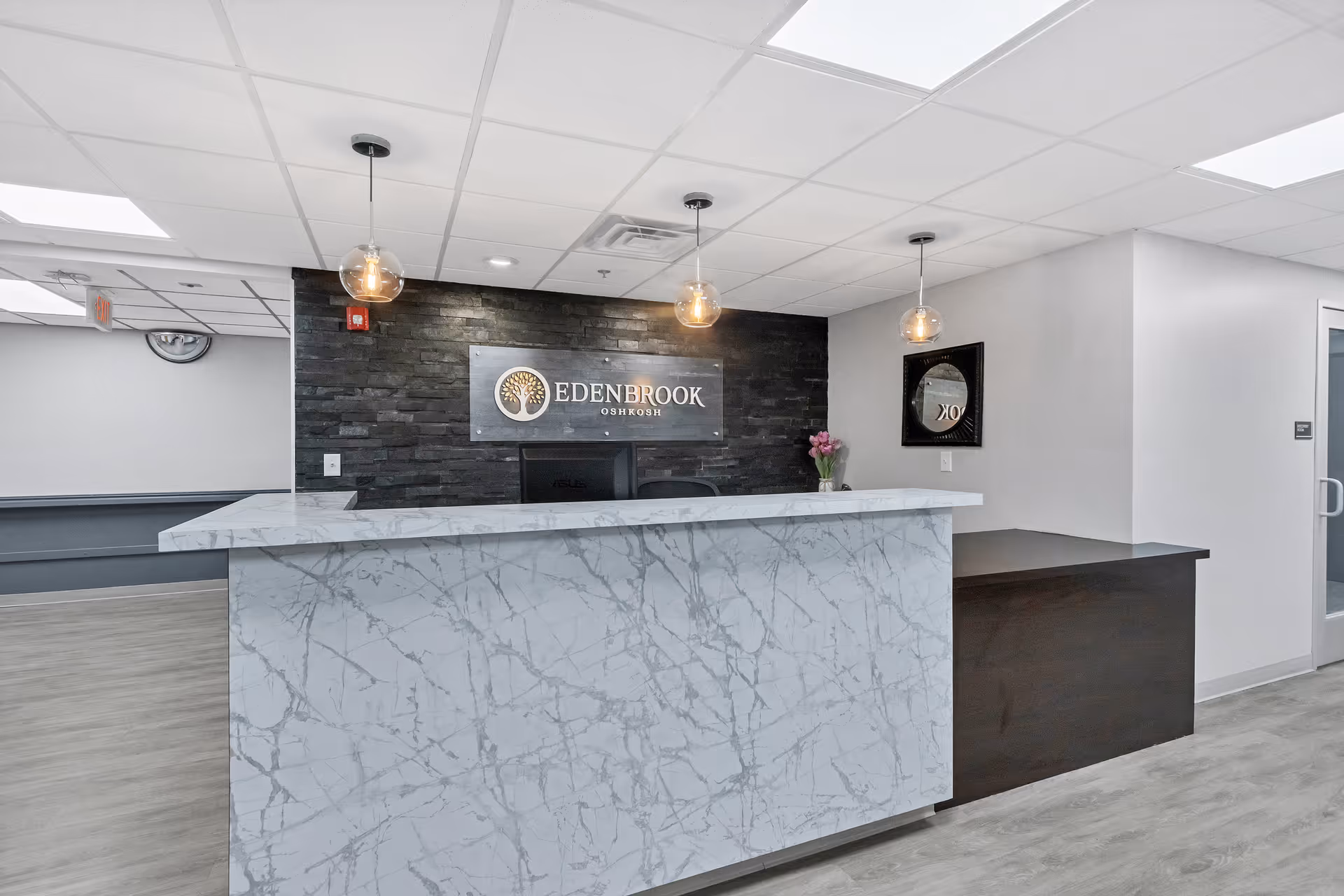 Reception desk area inside Edenbrook Oshkosh facility with a marble-patterned front counter, three hanging pendant lights, a black stone accent wall behind the desk with the Edenbrook Oshkosh logo, and a small vase with flowers on the counter.
