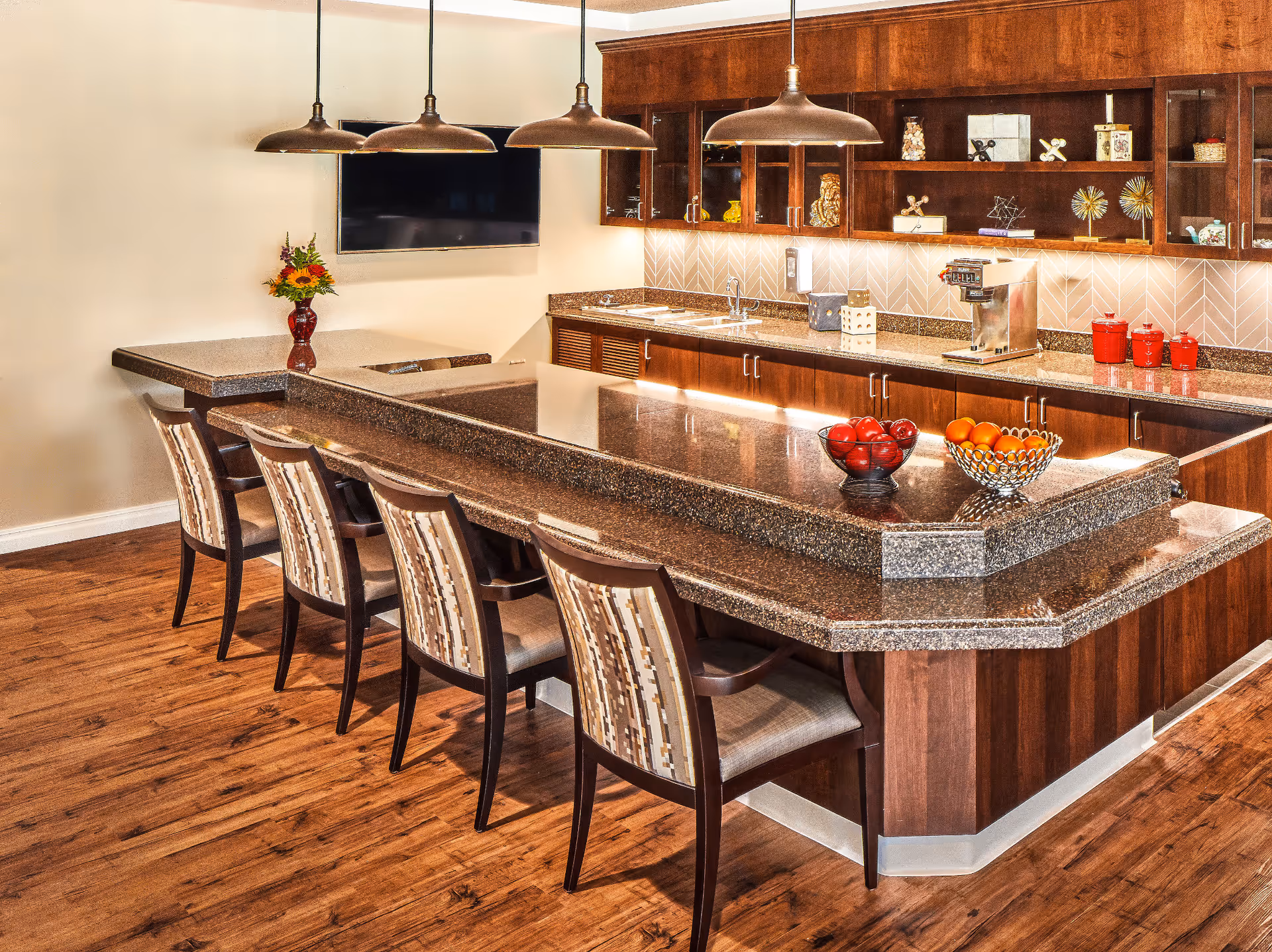 A modern kitchen area with a large granite countertop island surrounded by five cushioned chairs. The kitchen features wooden cabinets with glass doors, a coffee machine, decorative items on the shelves, and bowls of fruit on the counter. Pendant lights hang above the island, and a flat-screen TV is mounted on the wall. The floor is wooden, and there is a small table with a vase of flowers in the corner.