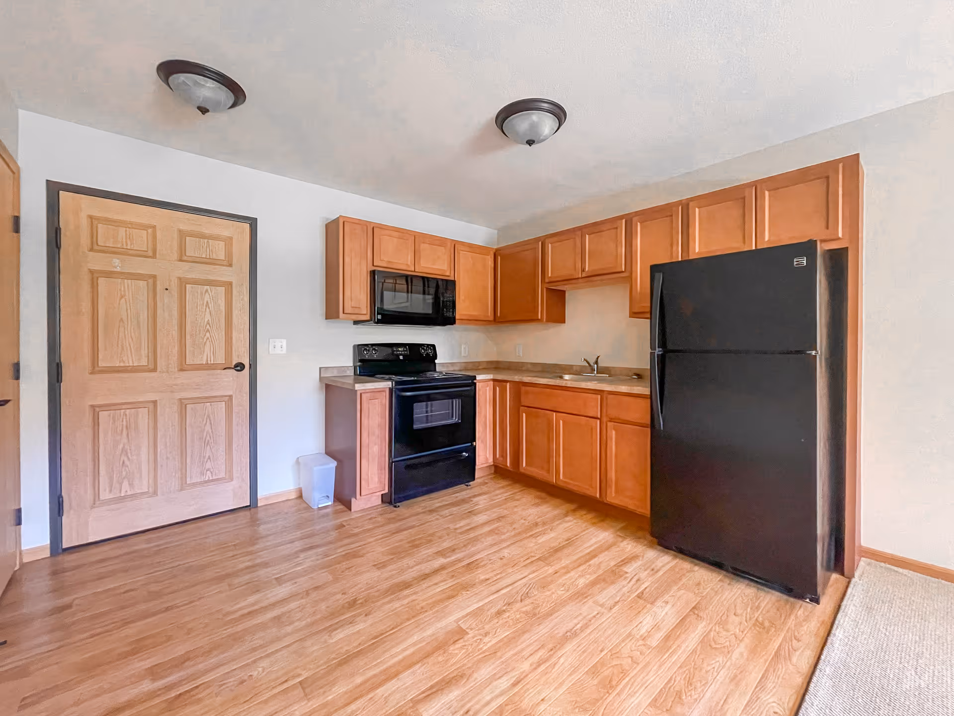 A small kitchen area with wooden cabinets, a black refrigerator, black microwave, and black stove. The floor is wood laminate, and there is a wooden door to the left. The walls are light-colored, and there are two ceiling light fixtures.
