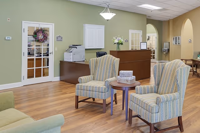 A reception area with two striped armchairs and a small round wooden table between them. Behind the chairs is a wooden reception desk with a vase of flowers on top. The walls are painted light green and beige, with a door featuring glass panes and a decorative wreath. The floor is wooden, and there is a printer on a side table near the reception desk.