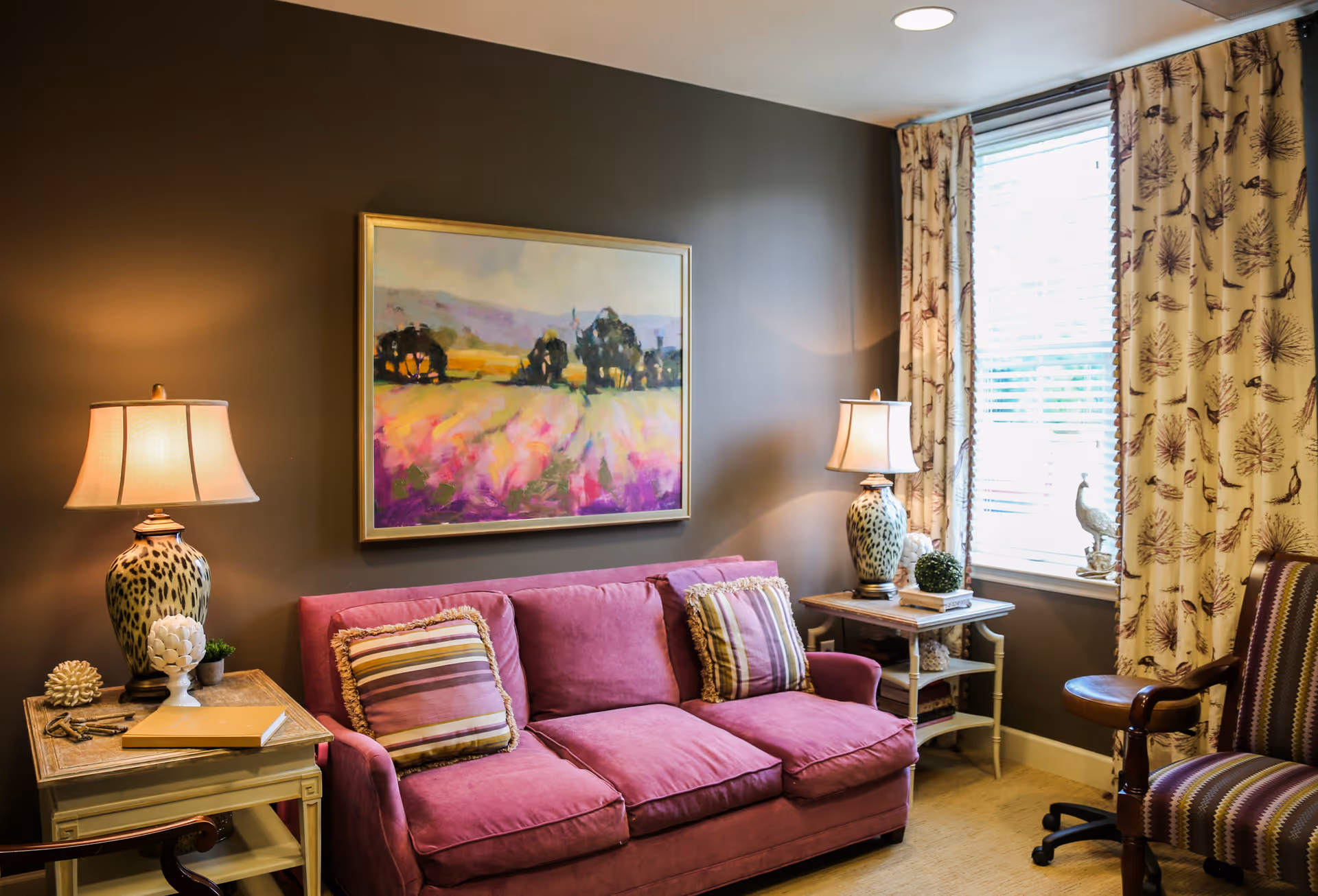 Cozy sitting room with a pink sofa, striped cushions, a framed painting above, two table lamps, patterned curtains and a window.