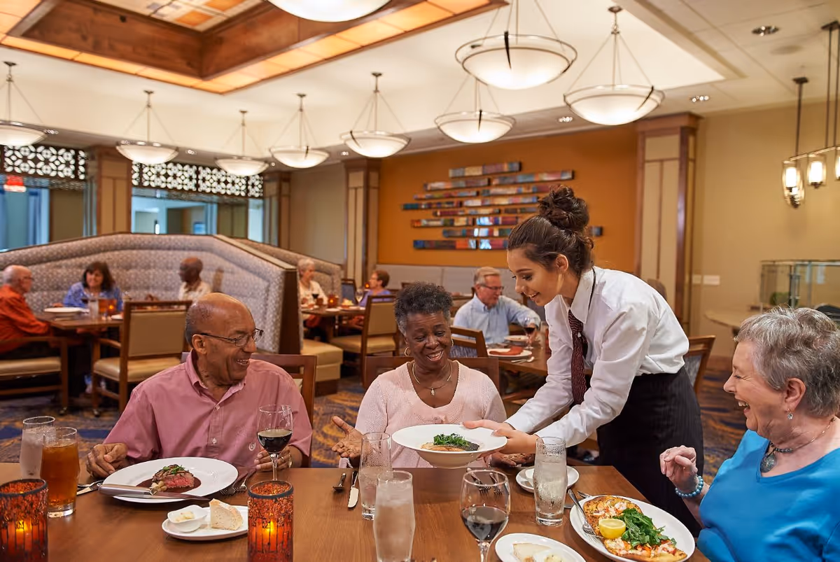 A waitress serving a dish to a smiling elderly woman seated at a dining table with two other elderly people in a well-lit restaurant. The table has plates of food, glasses of water, and wine. Other diners are visible in the background, seated in booths and chairs.
