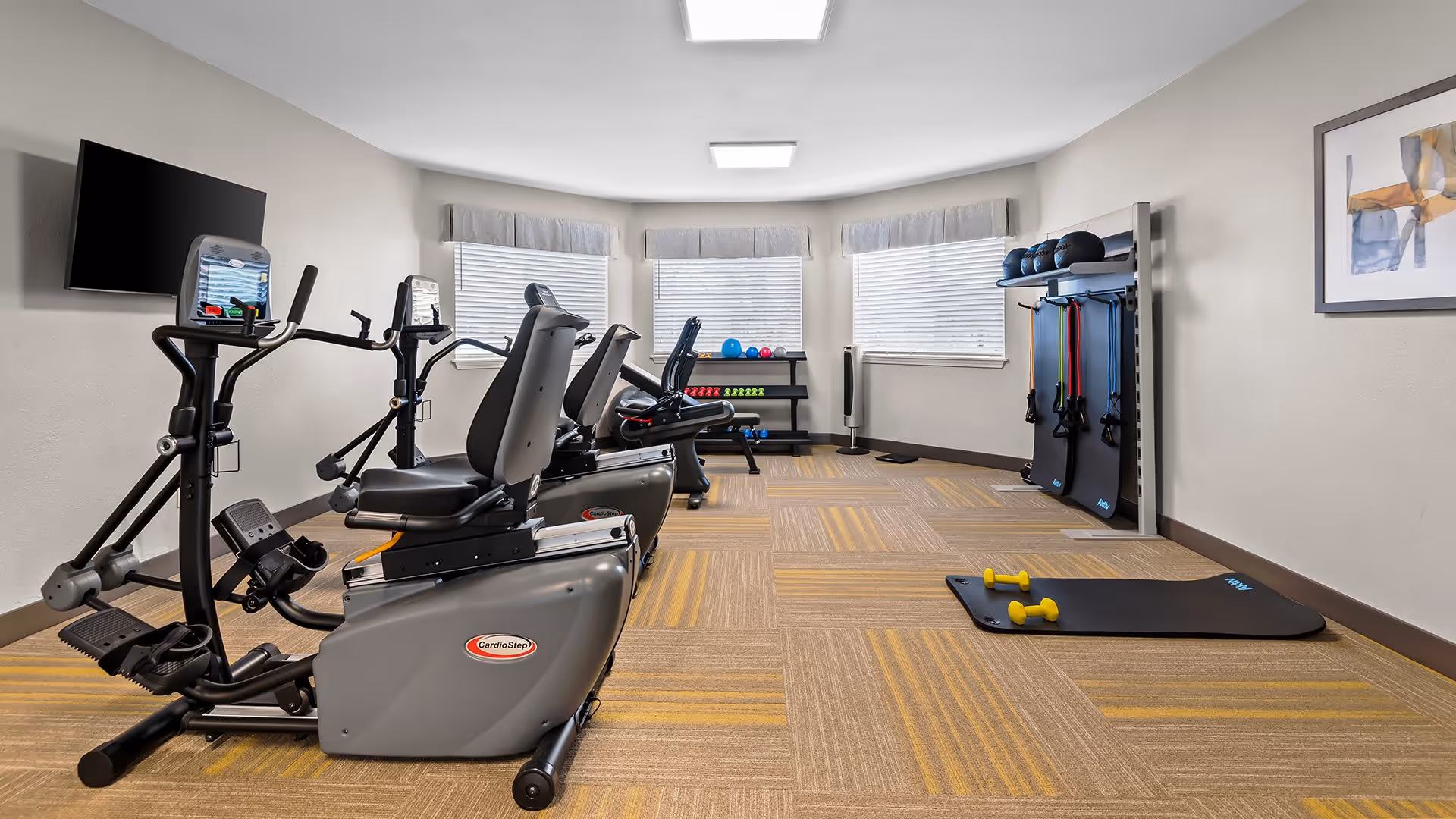 A small fitness room with exercise equipment including two recumbent cardio machines, a rack with various weights and exercise balls, resistance bands hanging on the wall, and two exercise mats with yellow dumbbells on the floor. The room has three windows with blinds and light-colored walls and carpeted floor with a yellow and brown pattern.