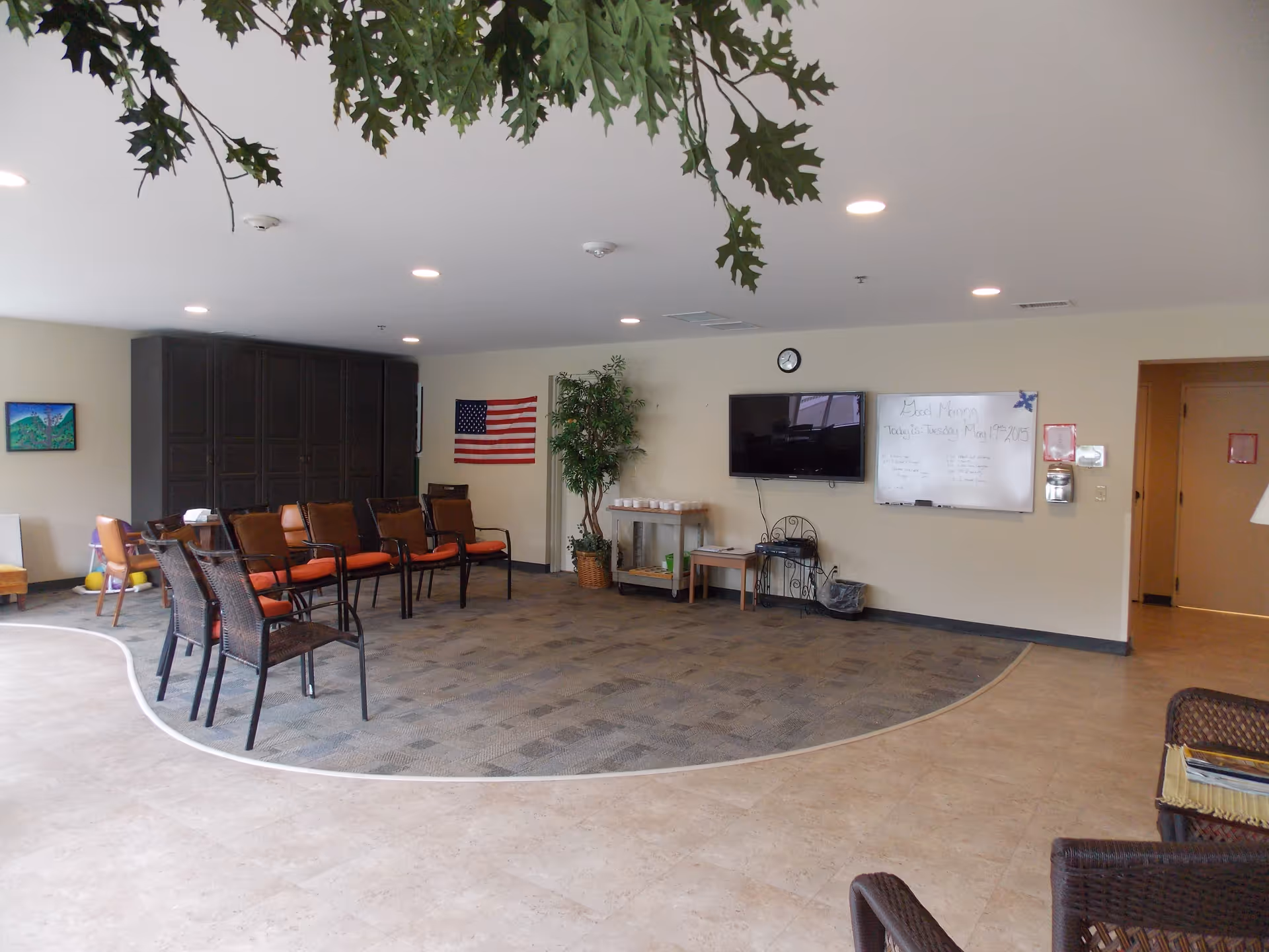 A spacious common area in a senior living facility with a semi-circle arrangement of chairs on a carpeted section of the floor. The room has beige walls, a mounted flat-screen TV, a whiteboard with notes, an American flag, a potted plant, and a few small tables. The ceiling has recessed lighting, and there is a partial view of wicker furniture in the foreground.