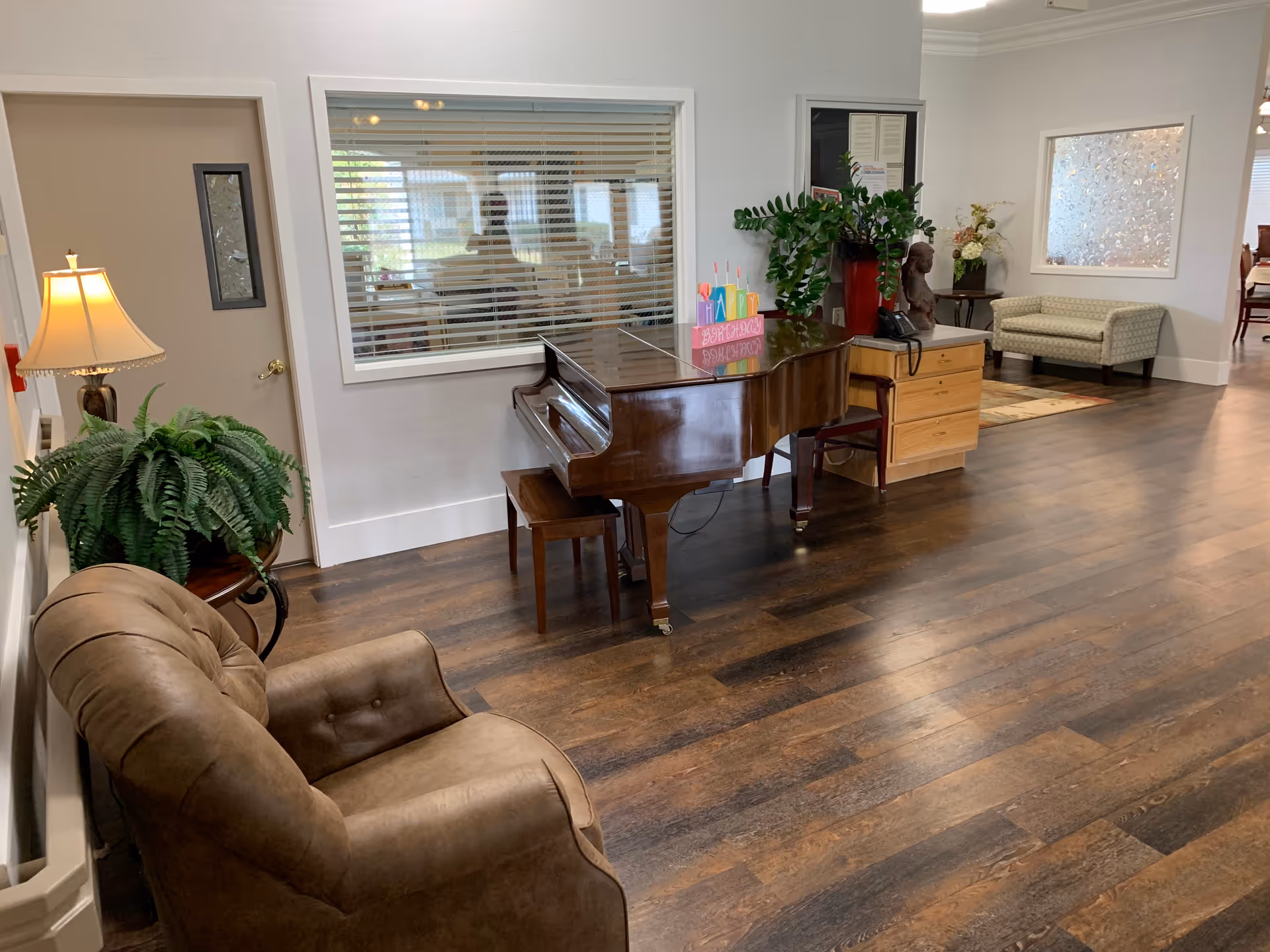 Assisted living common area with a grand piano, seating, plants and hardwood floors.