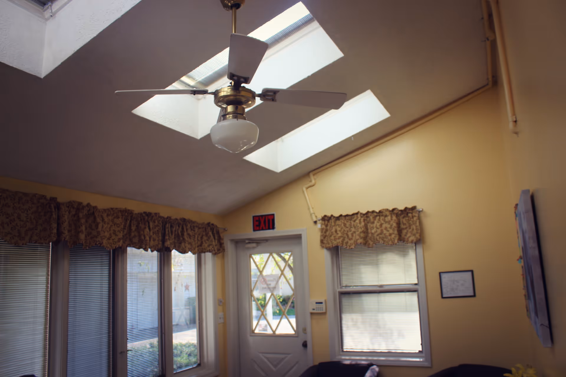 Interior view of a room with yellow walls, two skylights in the ceiling, a ceiling fan with a light fixture, windows with blinds and floral valances, a white door with a diamond-patterned window, and an exit sign above the door.