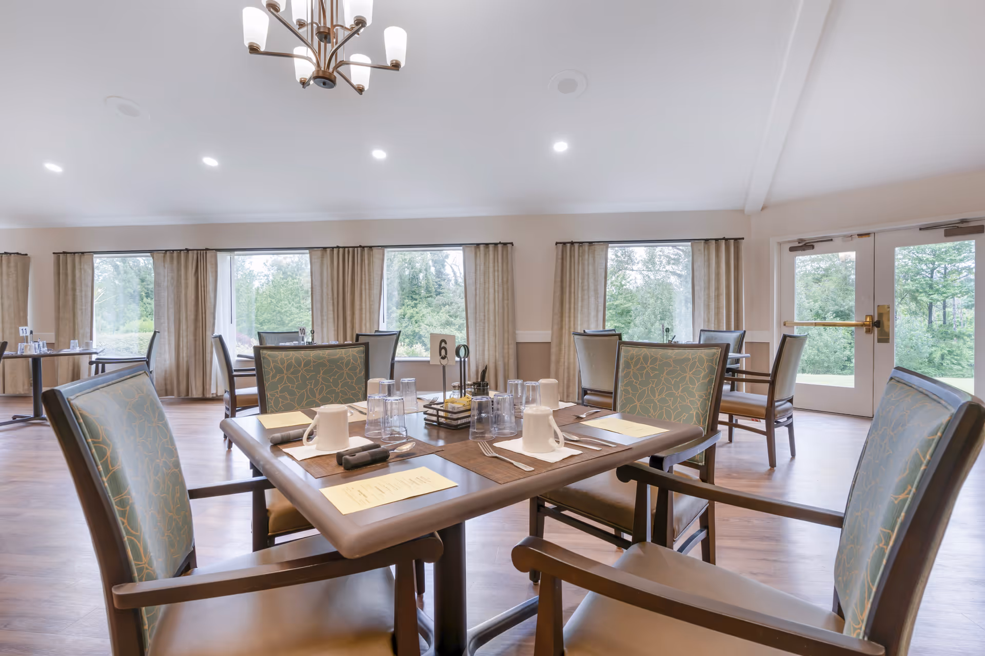 A bright dining room with several tables and chairs arranged neatly. Each table is set with menus, cups, glasses, and utensils. Large windows with beige curtains allow natural light to fill the room, and a chandelier hangs from the ceiling. Double glass doors lead outside to a green outdoor area.