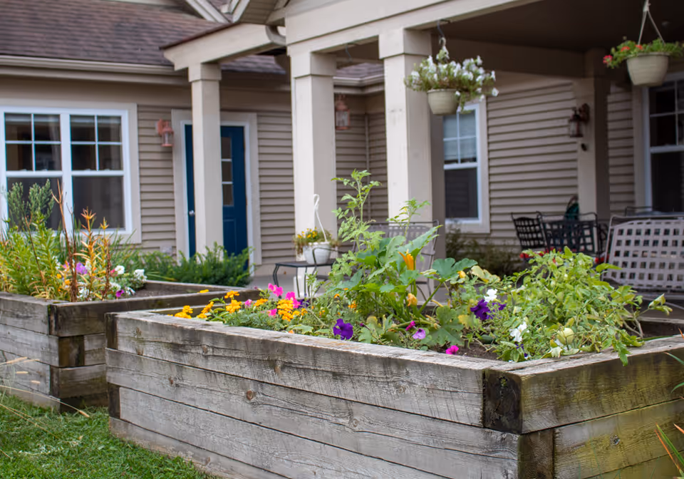 Raised wooden garden beds filled with various flowering plants and greenery in front of a beige building with white-framed windows and a blue door. There are hanging flower pots and outdoor seating visible under a covered porch area.
