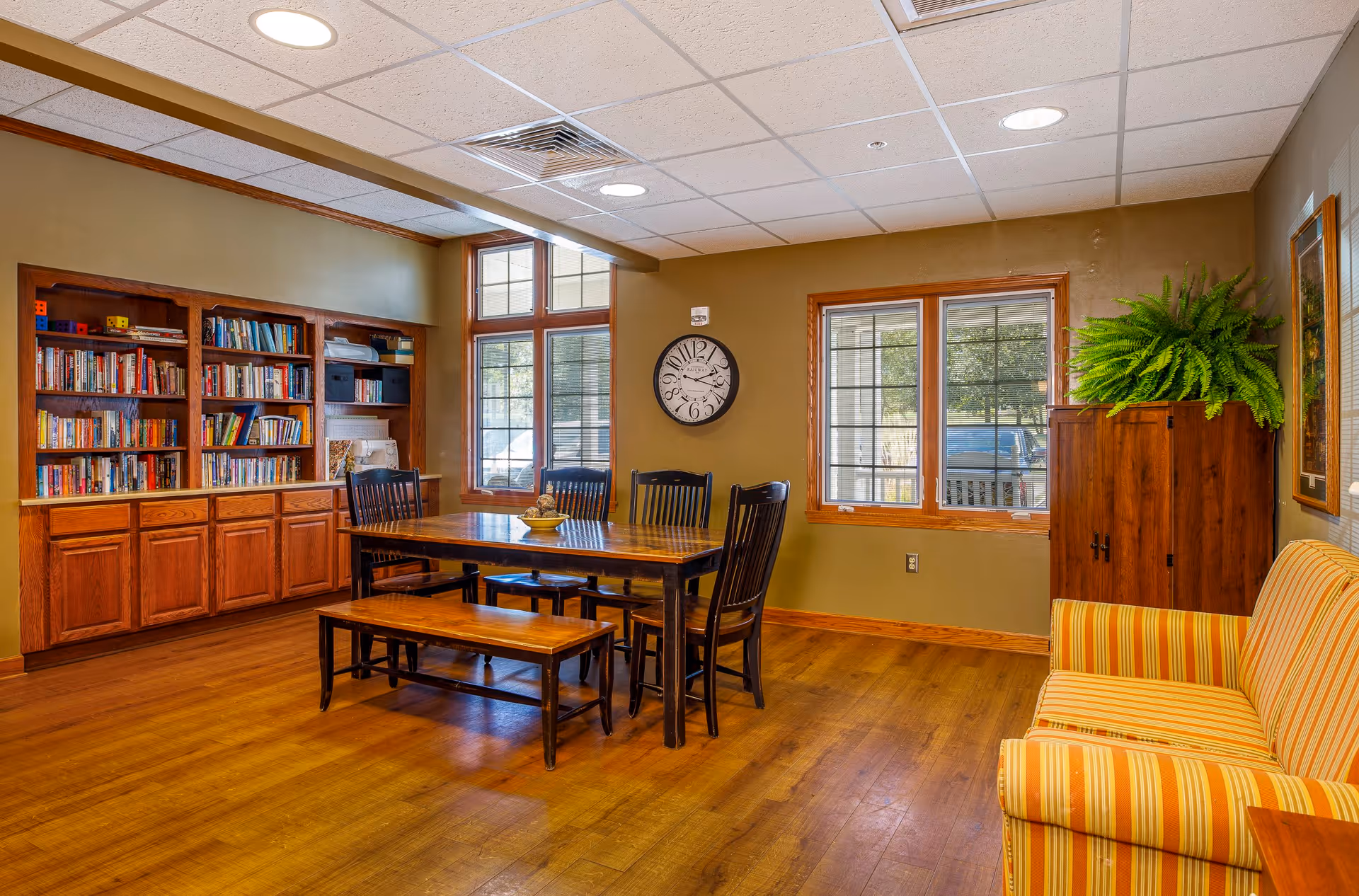 A cozy interior room with wooden flooring and beige walls featuring a wooden dining table with four chairs and a bench. There is a built-in wooden bookshelf filled with books on the left side, two large windows letting in natural light, a wall clock, a wooden cabinet with a green fern plant on top, and a striped yellow and orange sofa on the right.