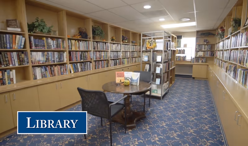 Interior view of a library room with wooden bookshelves filled with books along the walls. A round wooden table with two chairs is in the center, displaying a few books. The floor is covered with a patterned blue carpet, and the ceiling has recessed lighting.