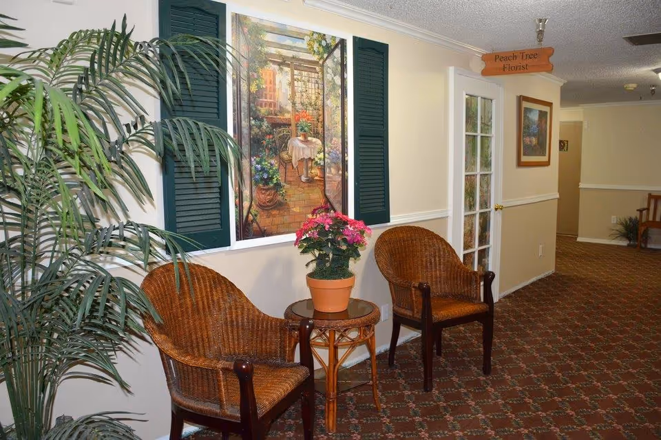 A hallway area in a senior living facility with two wicker chairs and a small round table with a potted pink flower. There is a large decorative wall painting framed by green shutters, a tall potted plant on the left, and a sign above a door that reads 'Peach Tree Florist'. The carpet has a patterned design and the walls are painted beige with white trim.