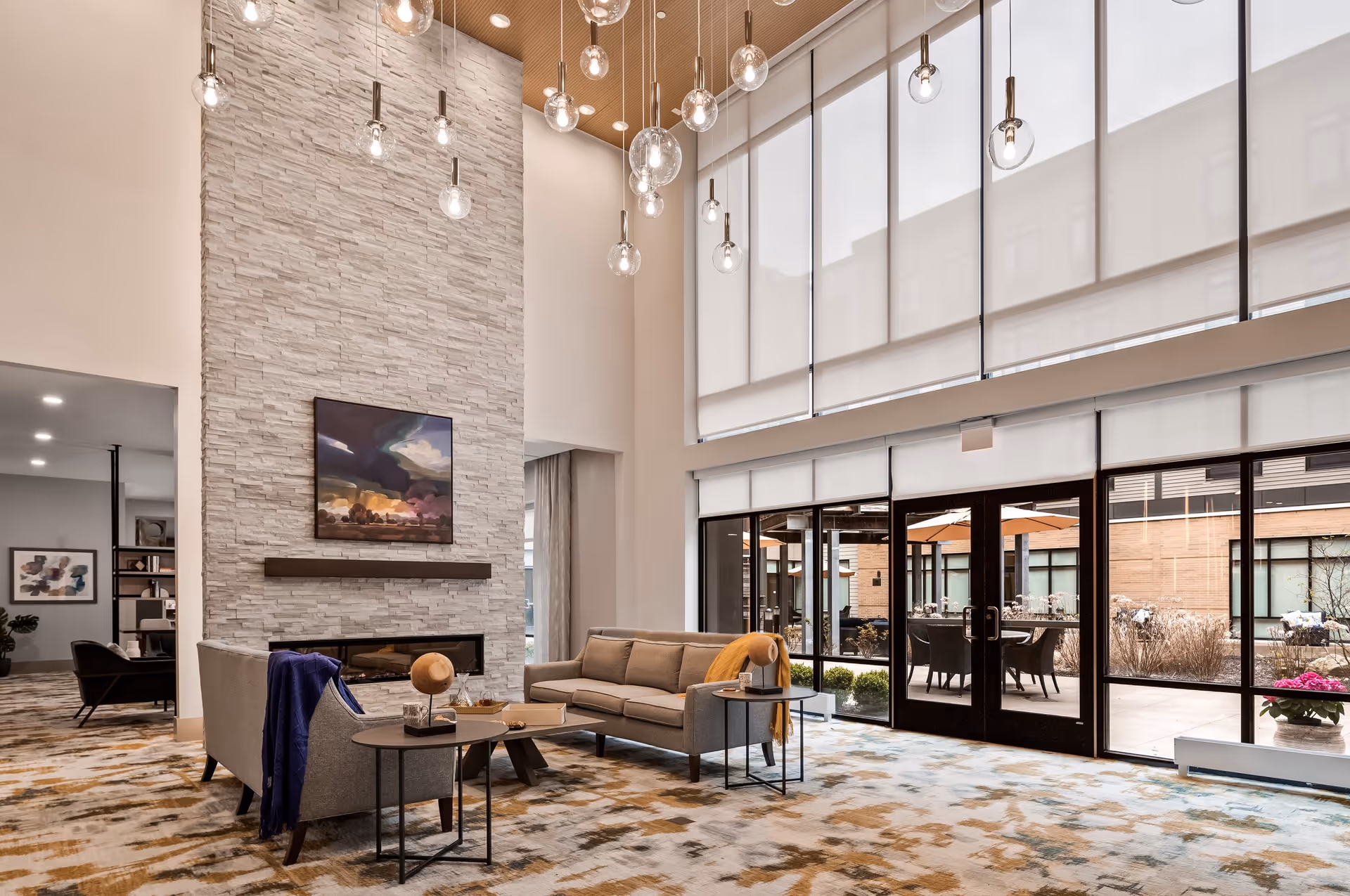 A spacious and modern living room area with high ceilings and large windows covered by white roller shades. The room features a tall stone fireplace with a colorful painting above it, two gray sofas arranged around a small coffee table, and multiple hanging pendant lights with exposed bulbs. Outside the glass doors, there is a patio area with tables and umbrellas.