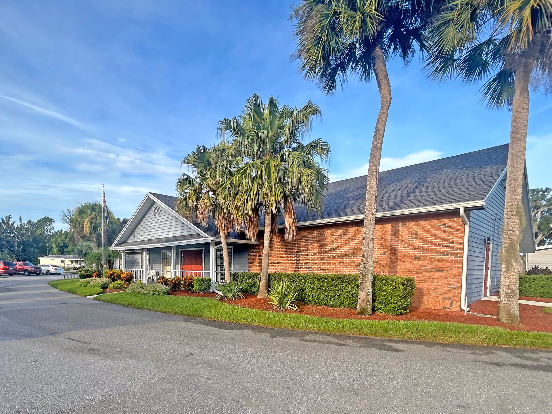 Single-story brick and siding retirement community building with a front porch, palm trees, and a parking area under a blue sky.