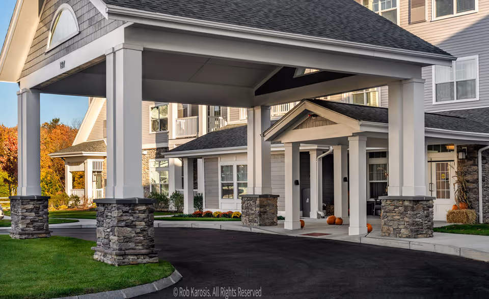 Entrance of a retirement community building with a covered driveway supported by white columns with stone bases. The building has beige siding and multiple windows, with pumpkins and hay bales placed near the entrance, suggesting a fall season.