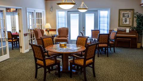 A cozy common area in a senior living facility with a round wooden table surrounded by six cushioned chairs in the foreground. In the background, there are two armchairs with a small table and lamp between them, a piano against the wall, framed artwork, and large windows with blinds letting in natural light. The room has carpeted floors and soft, warm lighting.
