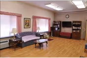 A living room with wooden flooring, a blue patterned sofa, a wooden coffee table, two windows with pink curtains, a side table with a lamp, a wall clock, a flat-screen TV mounted on the wall, and two wooden bookcases filled with books and decorative items.