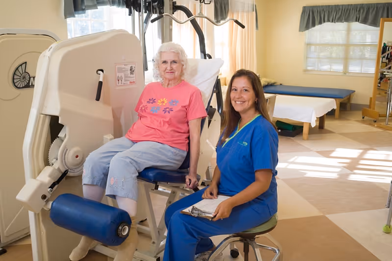 An elderly woman in a pink shirt and light blue pants is seated on a leg exercise machine in a rehabilitation room. A smiling female healthcare worker in blue scrubs sits beside her on a stool, holding a clipboard. The room is well-lit with large windows, exercise equipment, and therapy tables in the background.