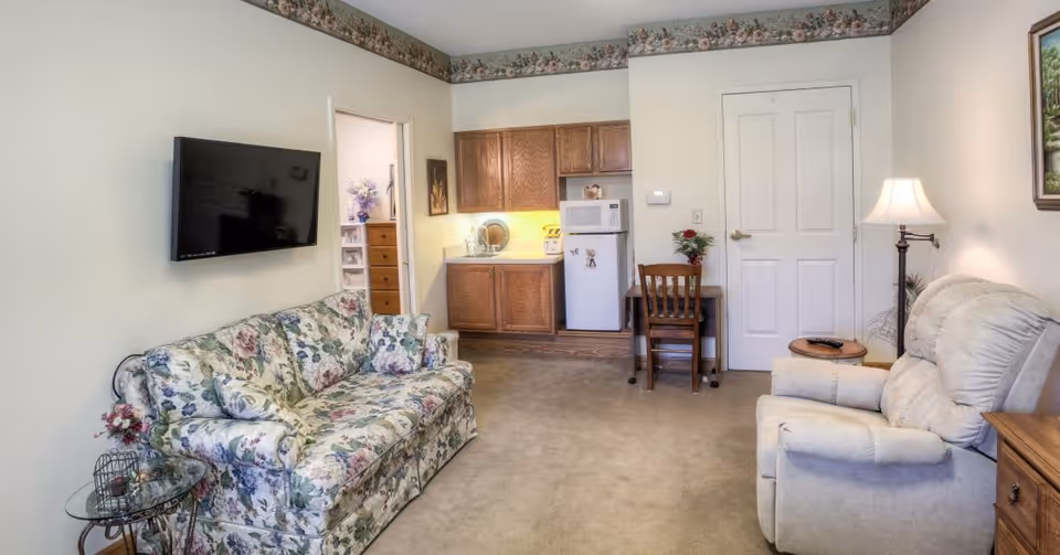 A cozy living room area with a floral patterned sofa and a beige recliner chair. A flat-screen TV is mounted on the wall above the sofa. There is a small glass side table with a decorative item next to the sofa. The room has a small kitchenette with wooden cabinets, a microwave, and a mini refrigerator. A wooden chair and desk are positioned near a closed white door. The walls are light-colored with a floral border near the ceiling, and a floor lamp is placed beside the recliner.