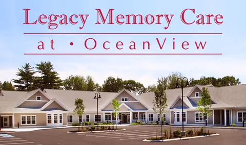 Exterior view of a single-story senior living facility building with multiple entrances, surrounded by small trees and a parking lot under a clear sky. The text 'Legacy Memory Care at Ocean View' is displayed prominently above the building.