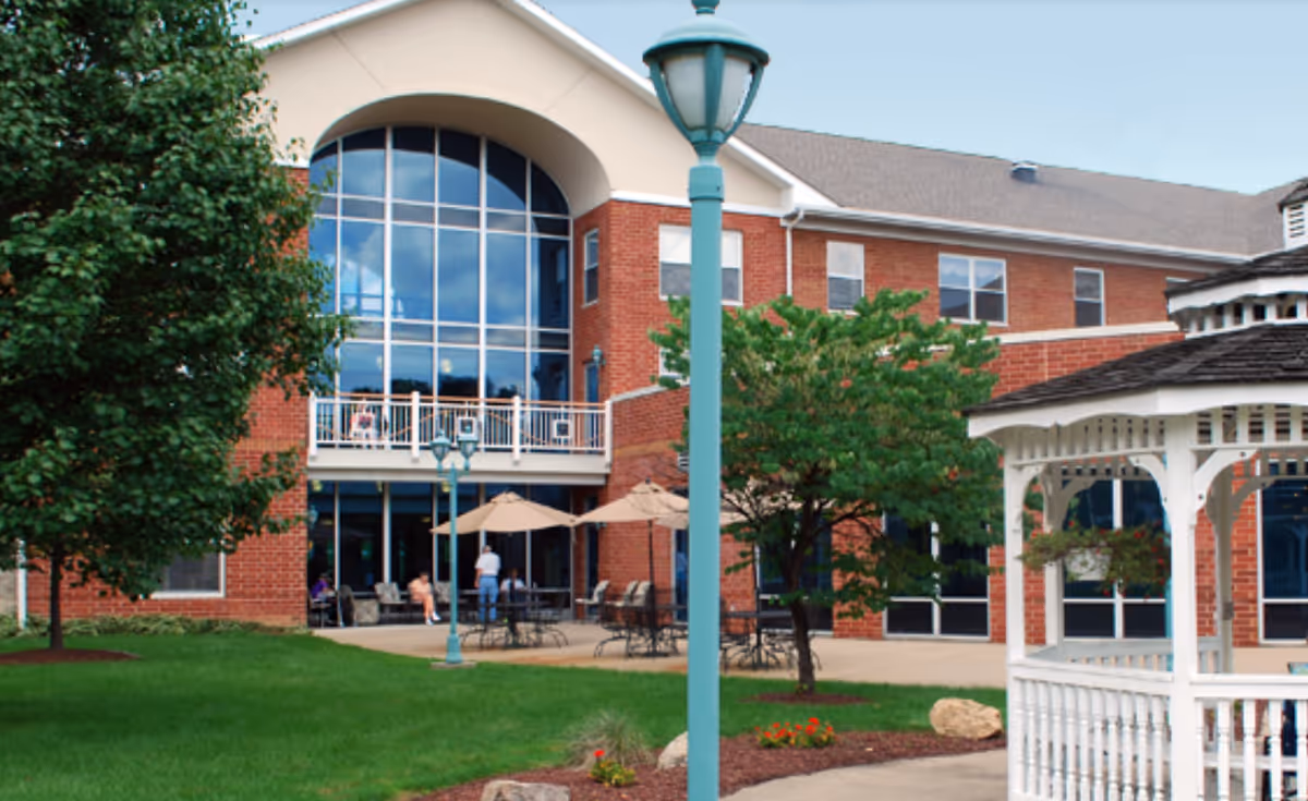 Outdoor view of a senior living facility named Brigham House featuring a red brick building with large arched windows, a patio area with tables and umbrellas, green trees, a white gazebo, and a green lawn.
