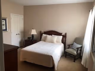 A bedroom with a neatly made bed featuring a wooden headboard, two white pillows, and a white quilt. There is a wooden dresser on the left side, a nightstand with a lamp and a small plant, and a cushioned chair with a pillow near a window with white curtains. The walls are painted beige and there is a closed double-door closet.