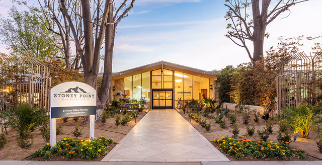 Exterior view of Stoney Point Healthcare Center at dusk, showing a modern building with large glass windows and a well-lit interior. A tiled walkway leads to the entrance, flanked by landscaped gardens with flowers and small shrubs. Tall trees and a metal fence are visible on either side of the walkway.